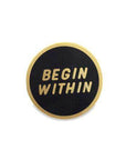 Black circle enamel pin with "begin within" in gold with gold border.