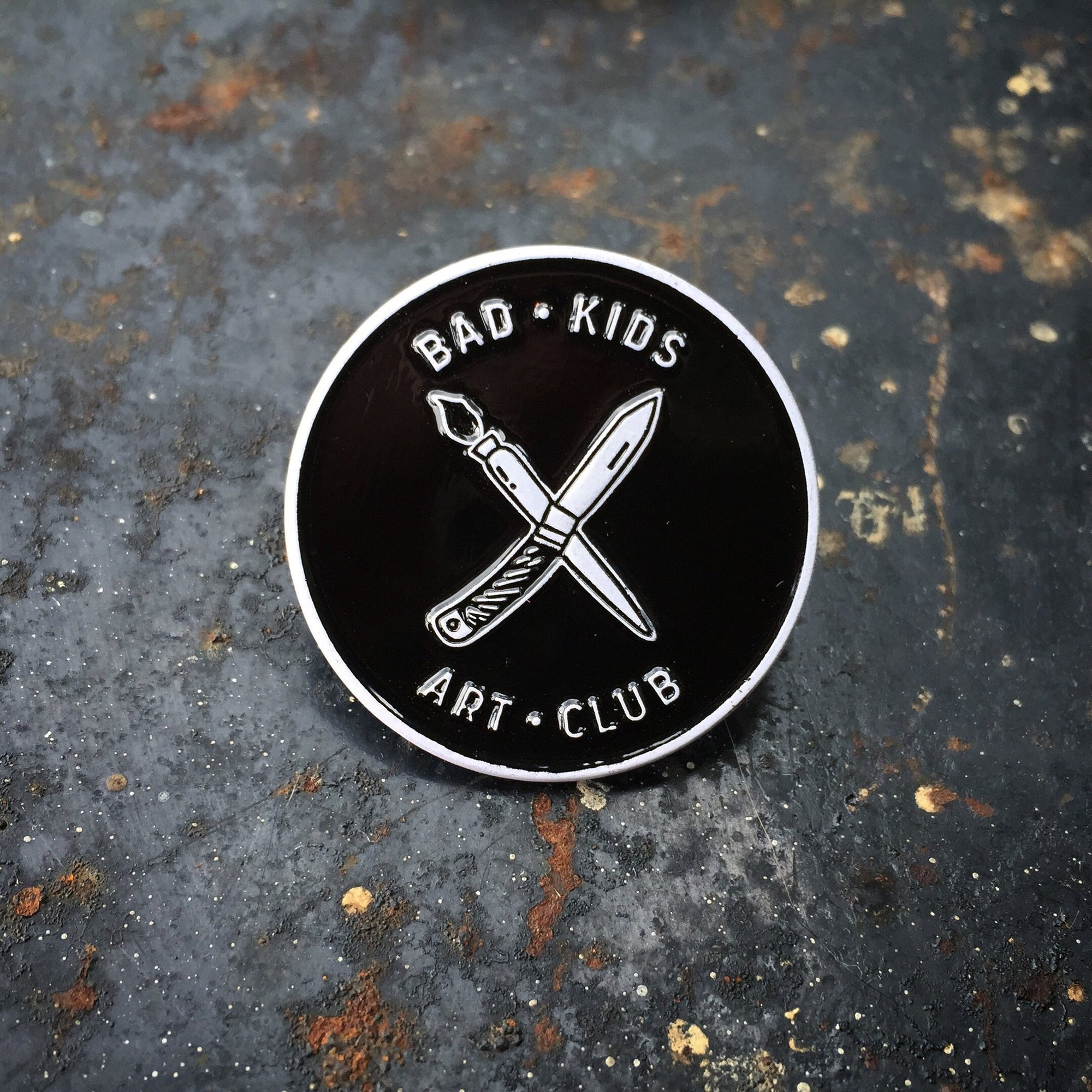 Circle enamel pin with &quot;x&quot; of paintbrush and knife with Bad Kids Art Club around it. By Print Ritual