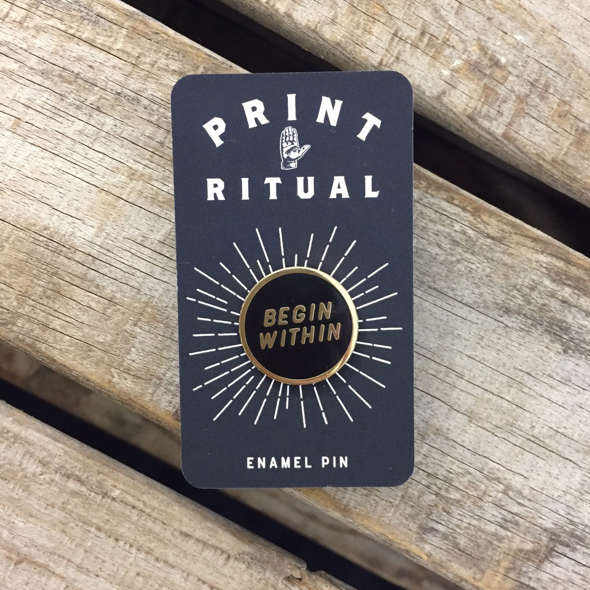 Black circle enamel pin with &quot;begin within&quot; in gold with gold border on print ritual card backing