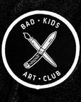 Circle iron on patch with "x" of paintbrush and knife with Bad Kids Art Club around it embroidered in white. Merrowed edge in white. By Print Ritual